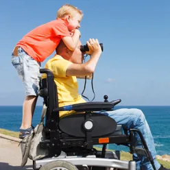 Mann i rullestol ser i kikket. Et barn står bak på rullestolen og smiler.