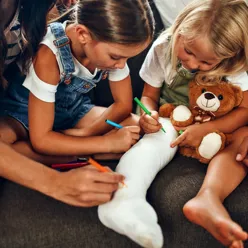 To barn som tegner og leker med en bamse. Det ene barnet har gips på benet
